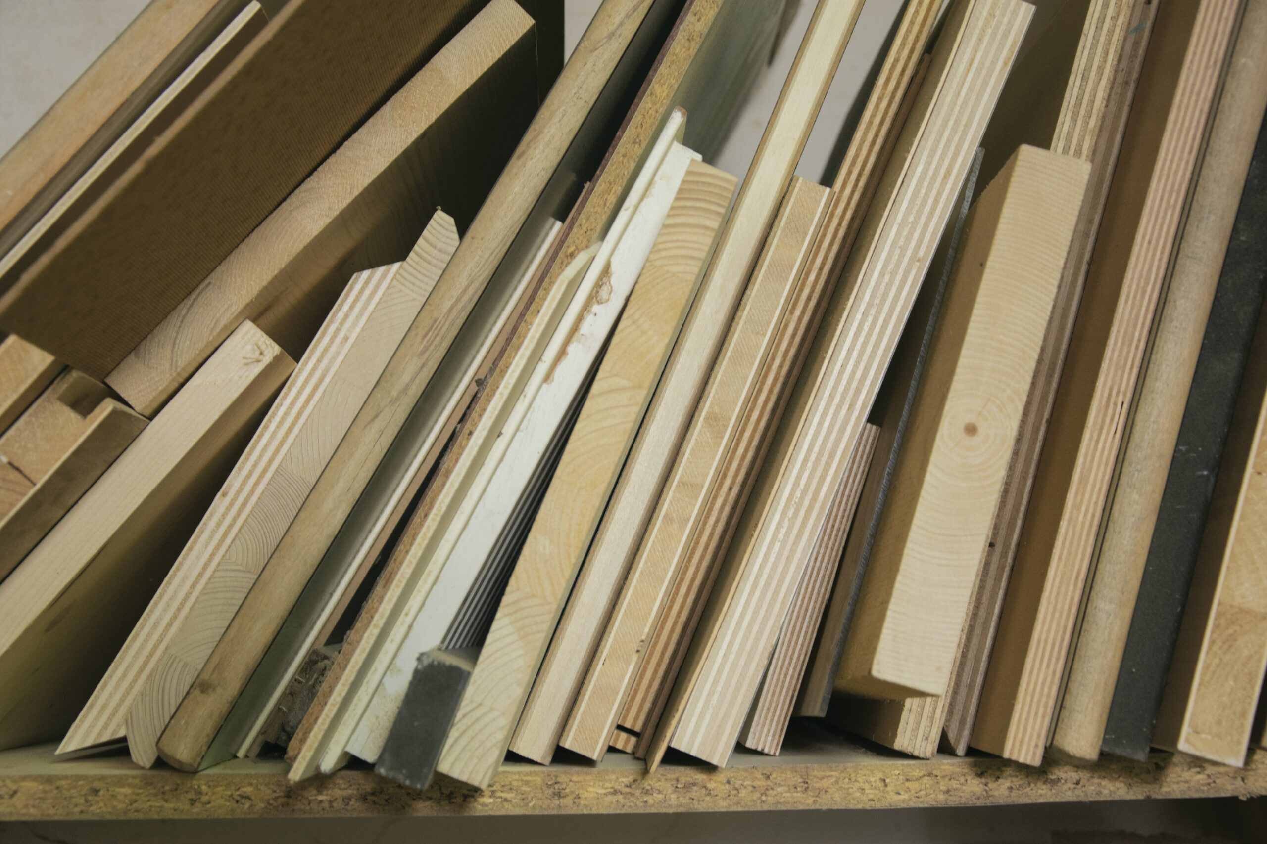 Stack of assorted wooden boards and plywood sheets stored vertically on a shelf in a workshop or lumber area.