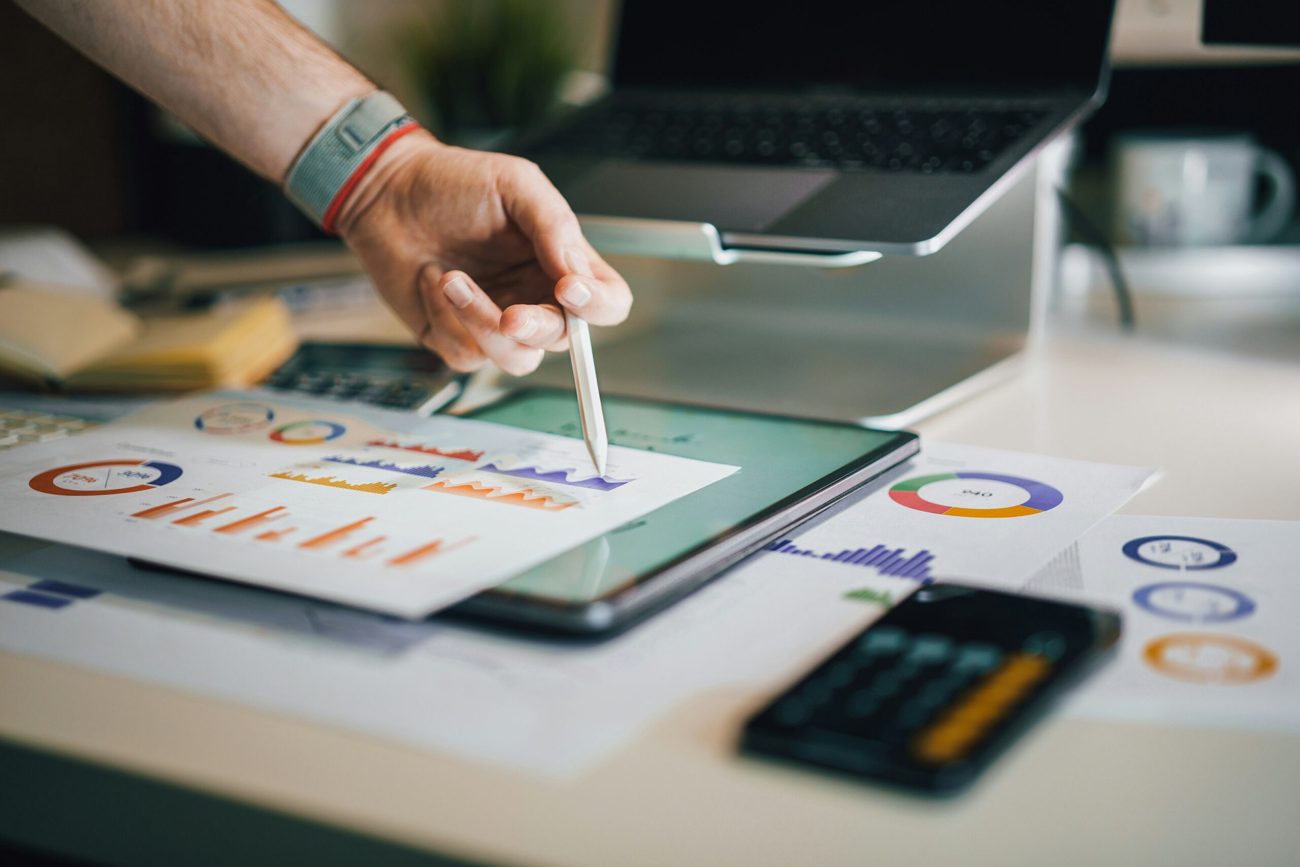 Hand using digital stylus on tablet displaying data analytics dashboard with colorful pie charts, bar graphs and KPI metrics for business performance tracking.