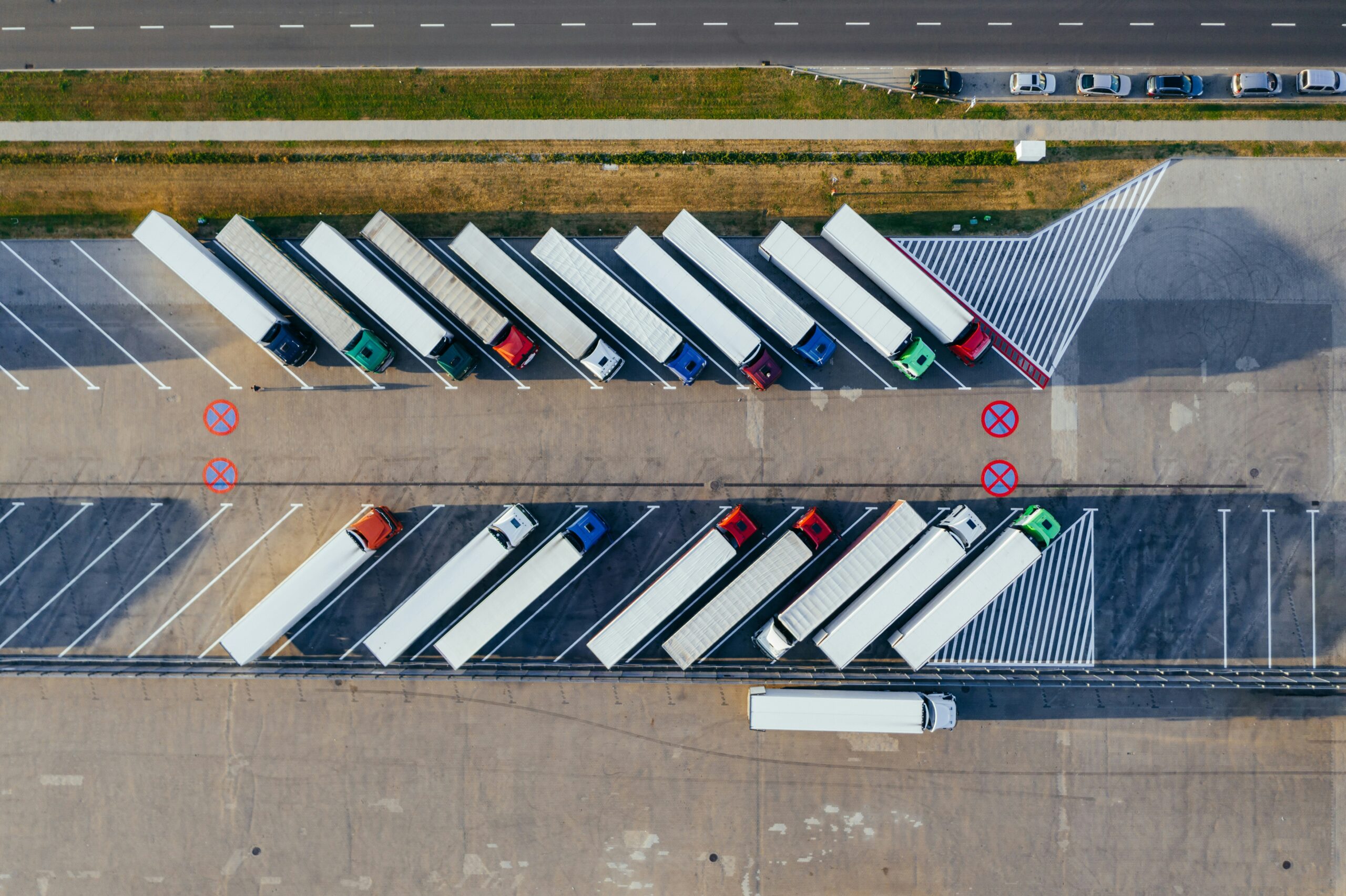 Aerial view of a parking lot filled with semi-trailer trucks parked in diagonal rows, adjacent to a road with cars and a sidewalk.