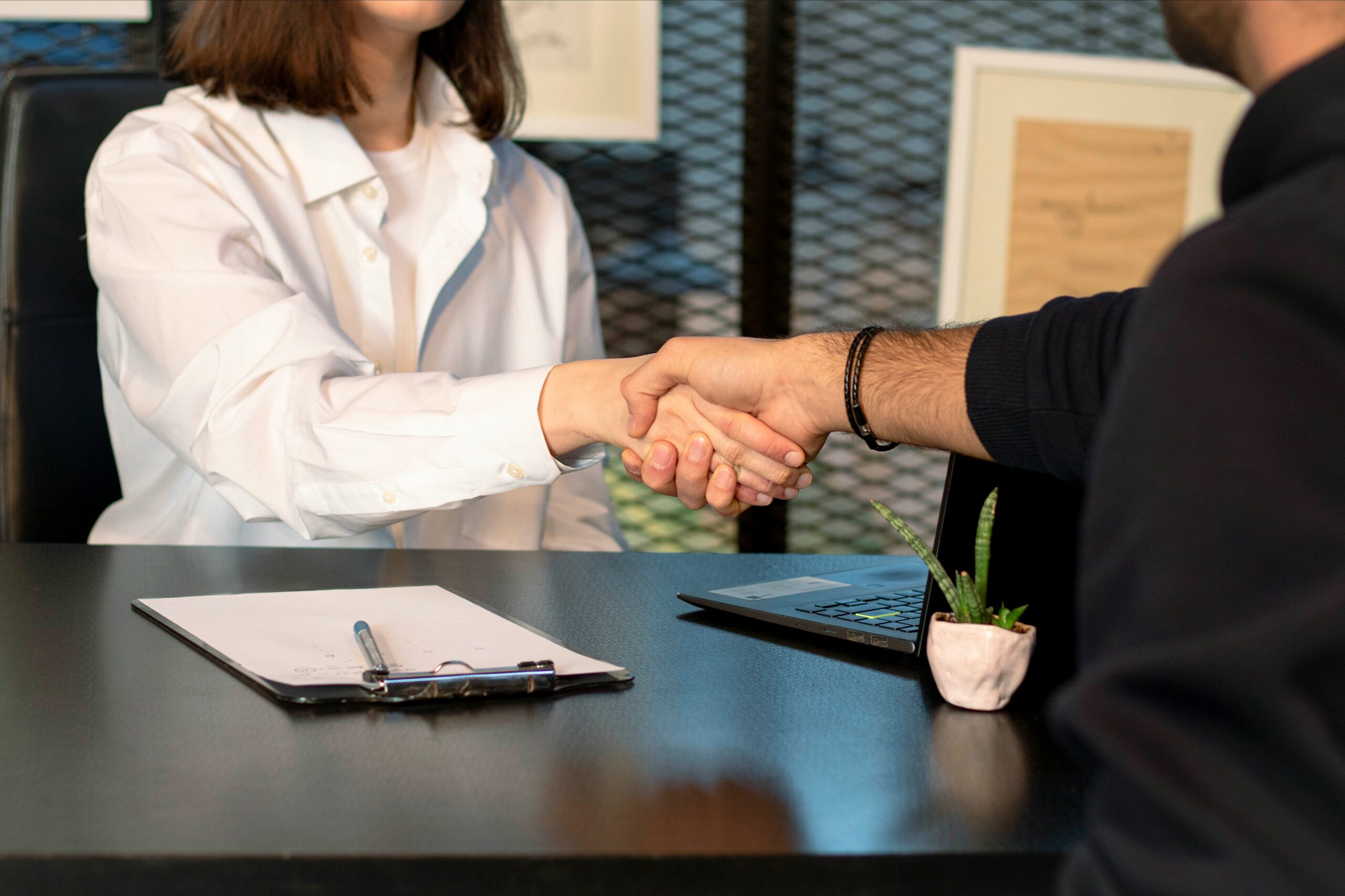 Two people shaking hands across a desk, symbolizing a successful business agreement or job interview, with a clipboard, laptop, and small plant on the table.