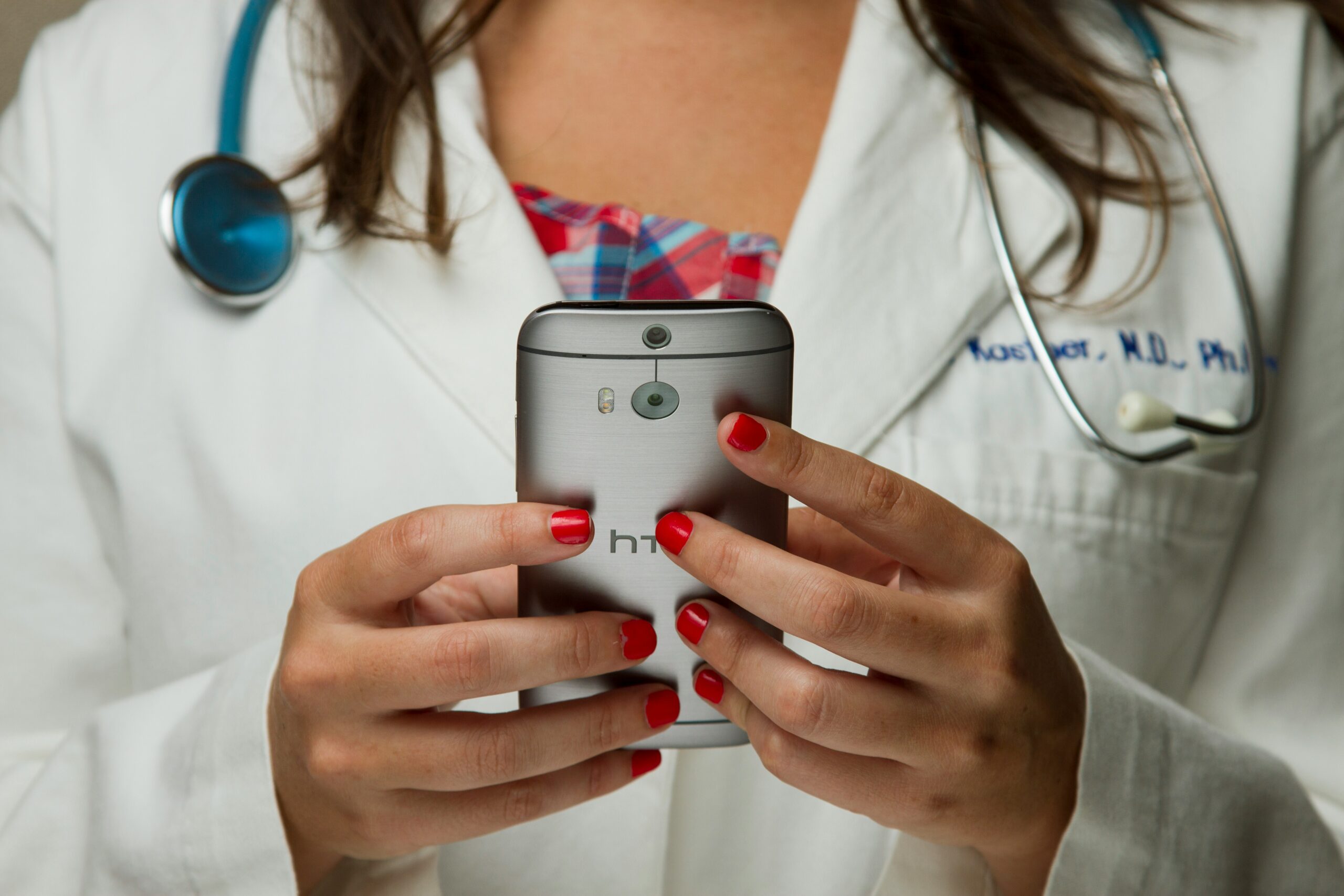 Close-up of a female doctor in a white coat using a smartphone, with a stethoscope around her neck and red nail polish on her fingers.