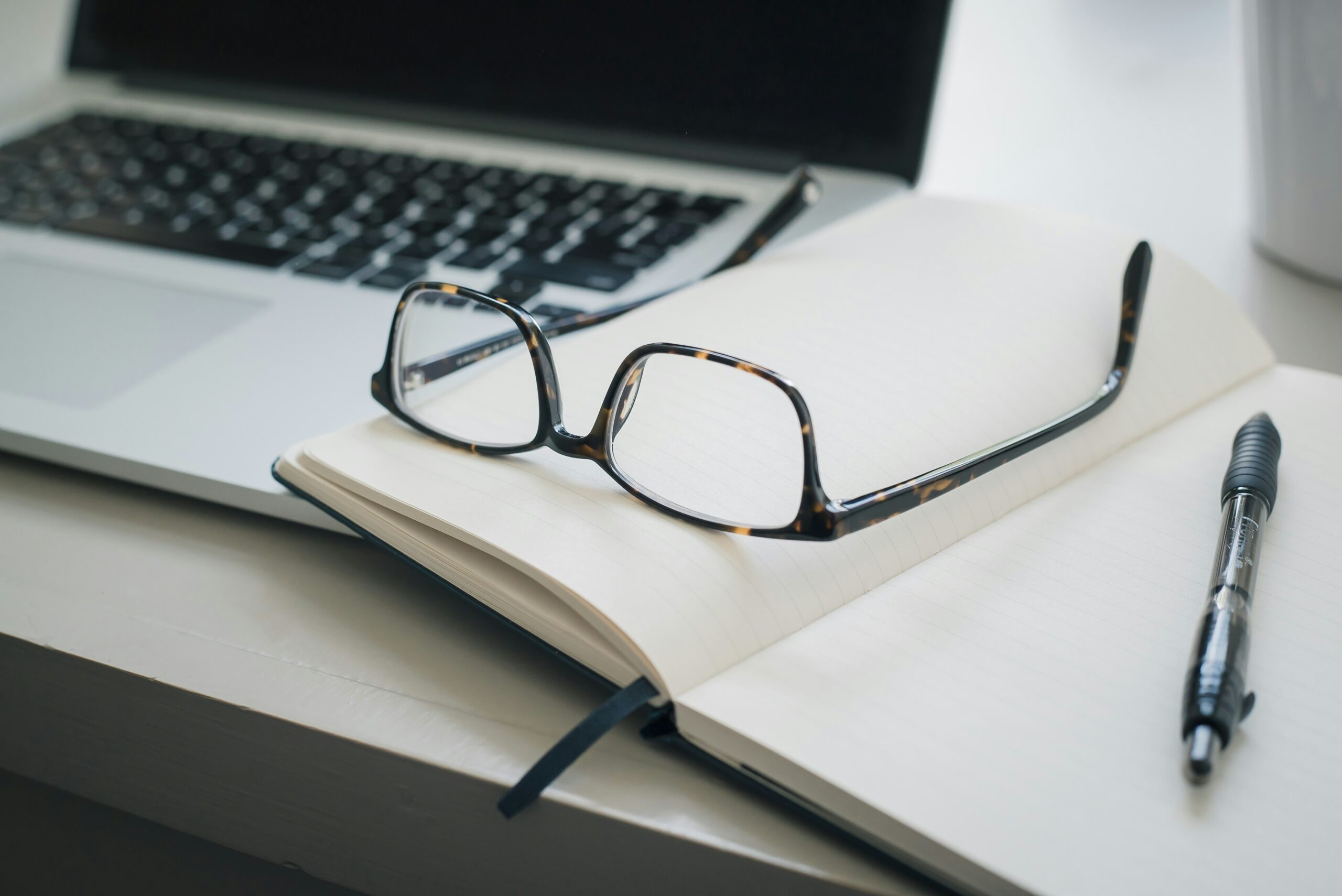 Eyeglasses resting on an open lined notebook beside a pen and a laptop on a white desk, suggesting a workspace or study setting.