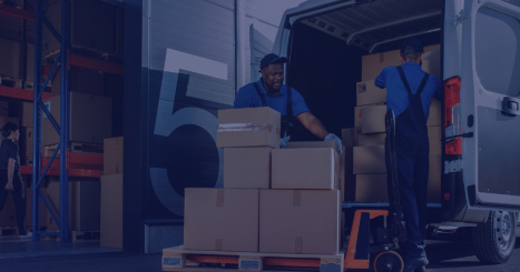 Two warehouse workers in blue uniforms load cardboard boxes from a delivery van onto a pallet inside a warehouse.